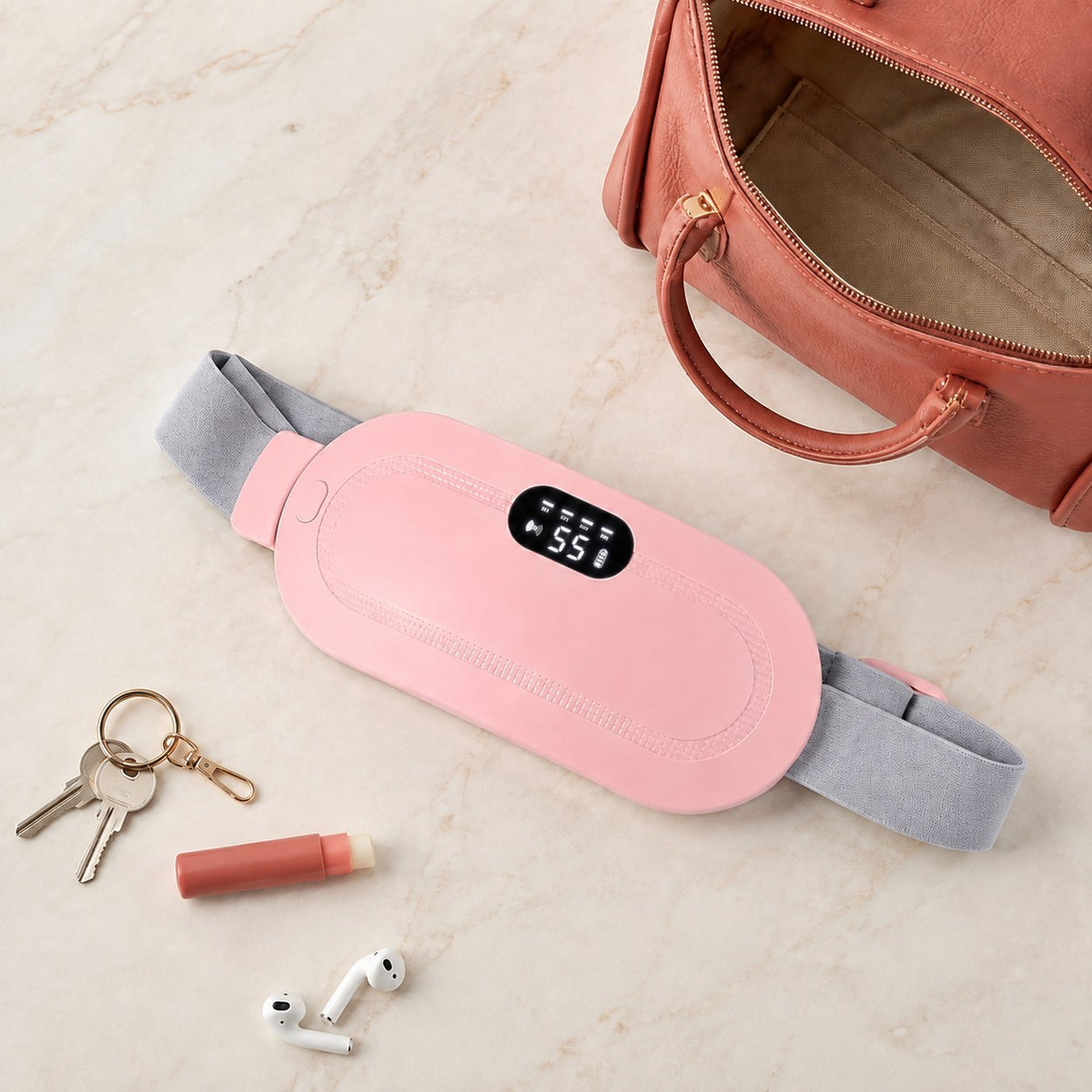Ceinture chauffante CHALYNE rose posée sur du marbre à côté d'un sac à main, de clés et d'écouteurs